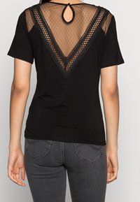 Kobieta w czarnym topie z krótkim rękawem, z przezroczastym, w kształcie litery V, koronkowym tyłem oraz w ciemnoszarych dopasowanych dżinsach, stojąca na jednolitym tle.