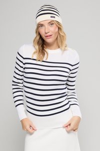 Blonde vrouw in een witte en navy gestreepte lange mouwen sweater en een bijpassende beanie, die de onderkant van haar sweater aanpast, op een neutrale grijze achtergrond.