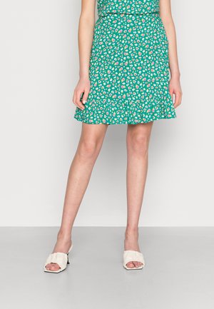 Saia verde com estampado floral e bainha rodada, apresentando flores brancas e rosas. Combinada com mules abertas de cor creme, exibindo um design com babados.