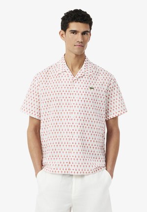 Hombre con camisa blanca de manga corta y botones, con patrón de conos de helado naranja, y pantalones blancos, con las manos en los bolsillos, de pie frente a un fondo blanco.