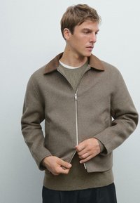 Brown wool zip-up jacket with a brown collar, fitted sleeves, and ribbed waistband. Underneath, a light green knitted sweater is visible.