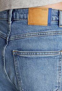 Jeans de denim azul claro con un parche de cuero en la cinturilla trasera con texto en relieve. Costuras visibles y bolsillos traseros.