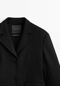 Manteau en laine noire avec col cranté, deux boutons et poches latérales. Texture unie, détails minimaux, et une étiquette visible à l'intérieur du col.