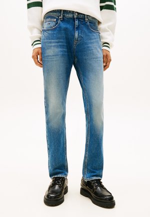 Mies, joka on pukeutunut vaaleansinisiin suoraan leikattuihin farkkuihin, joissa on lievää haalistumaa, mustiin nauhallisiin kenkiin ja valkoiseen neulepuseroon, jossa on vihreät raidat hihoissa.