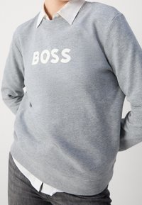 Sweatshirt gris en tissu doux, arborant un logo "BOSS" blanc en évidence sur la poitrine, associé à une chemise blanche à col en dessous.