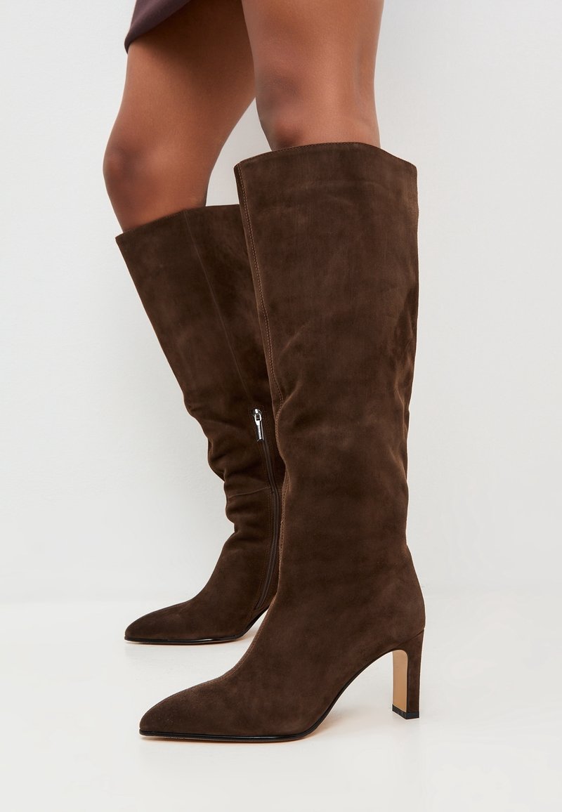 Botas altas hasta la rodilla de ante marrón con punta puntiaguda, tacón alto y delgado, y cremallera lateral. El material tiene una textura suave, proporcionando una apariencia lisa.