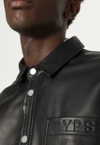 Giacca in pelle nera con design a bottoni, colletto e tasca frontale testurizzata con dettaglio in rilievo "YPS". Dettagli in metallo argentato.