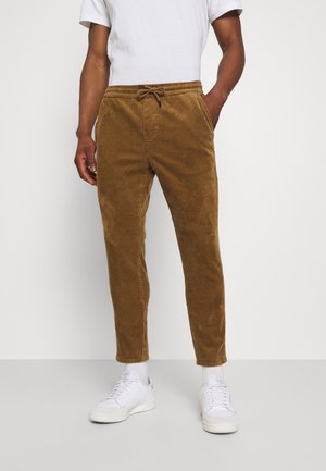 Pantalons en velours côtelé marron à coupe fuselée, avec une taille élastique, un cordon de serrage et des poches à l'avant, portés avec des baskets blanches.