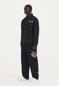 Giubbotto di jeans neri con chiusura a bottoni, tasche sul petto e logo ricamato bianco. Pantaloni neri con design ricamato colorato, abbinati a sneaker bianche.