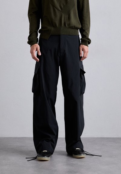 Pantaloni cargo negri cu o croială largă, prevăzuți cu buzunare laterale și șnururi la tiv, asortați cu un pulover tricotat de culoare verde închis.