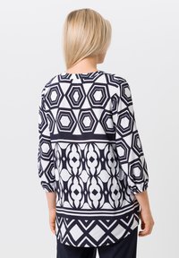 Blouse à motifs noir et blanc avec des manches trois-quarts, présentant des formes géométriques et un ourlet arrondi. Tissu lisse avec un léger éclat.