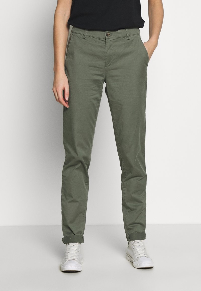 Personne portant un chino vert olive retroussé, une chemise noire et des baskets blanches, debout avec une main dans la poche.