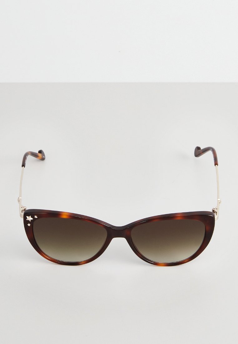 Des lunettes de soleil en écaille de tortue en forme de chat avec des branches en métal doré et des verres dégradés bruns sur un fond neutre.