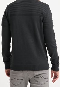 Schwarzer Pullover mit strukturierten horizontalen Streifen im oberen Rückenbereich sowie gerippten Bündchen und Saum. Kombiniert mit hellgrauen Jeans.