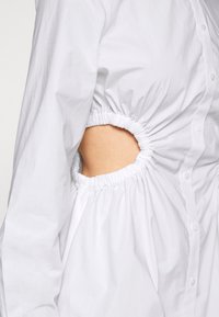 Vit skjorta med knappar, med insydda sidor och utskärningar under armarna. Lätt tyg, slät textur och mjuk midjedetalj.