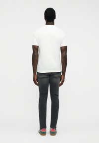 Hombre de piel oscura vistiendo una camisa blanca de manga corta lisa, jeans negros descoloridos y zapatillas verdes con calcetas rosas, de pie de espaldas.