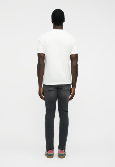 Άνδρας με σκούρο δέρμα που φοράει απλό λευκό κοντομάνικο πουκάμισο, ξεθωριασμένο μαύρο τζιν και πράσινα αθλητικά παπούτσια με ροζ κάλτσες, στέκεται με την πλάτη προς τα εμάς.