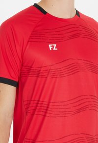 Punainen urheilupaita lyhyillä hihoilla, mustat yksityiskohdat kauluksessa, aaltoileva kuvio tummemmassa sävyssä, "FZ"-logo rinnassa.