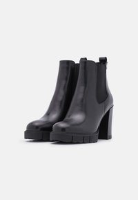 Bottes autour de la cheville en cuir noir avec une finition brillante, des panneaux latéraux texturés et des talons blocs épais. Comprend des tirettes pour un enfilage facile.