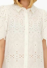 Camicia bianca realizzata in tessuto leggero, con motivi in broderie anglaise, frontale con bottoni e maniche corte a sbuffo.