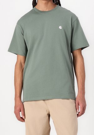 Hombre con camiseta Carhartt verde salvia y pantalones beige, de pie frente a un fondo blanco liso.