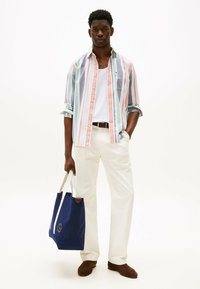 Gestreifte Hemdbluse in Pastellfarben, weißes Tanktop, cremefarbene Hose und dunkelbraune Slipper. Blaues Tote Bag mit Logo.