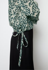 Widok z boku osoby w zielonej bluzce w kropki związanej w talii na czarnej spódnicy, na jednolitym tle.