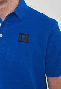 Polo shirt blu realizzato in tessuto testurizzato, con colletto, pattina con tre bottoni e un'etichetta quadrata nera con un logo.