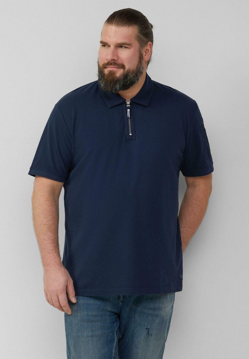 Granatowa koszulka polo z zamkiem błyskawicznym przy kołnierzy, krótkie rękawy i jednolity wzór. Noszona z niebieskimi dżinsami o luźnym kroju.