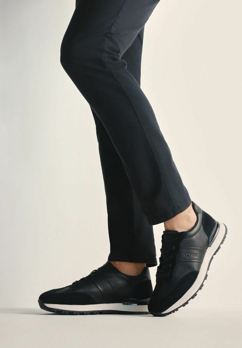 Czarne buty sportowe wykonane z połączenia skóry i zamszu, z białą podeszwą i teksturowanym wykończeniem, noszone z dopasowanymi spodniami.