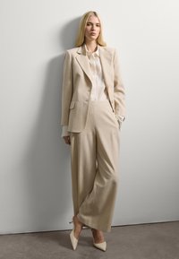 Vrouw staat tegen een witte muur gekleed in een beige pak met wijde broekspijpen, bijpassende blazer, witte blouse en crèmekleurige puntige hakken.