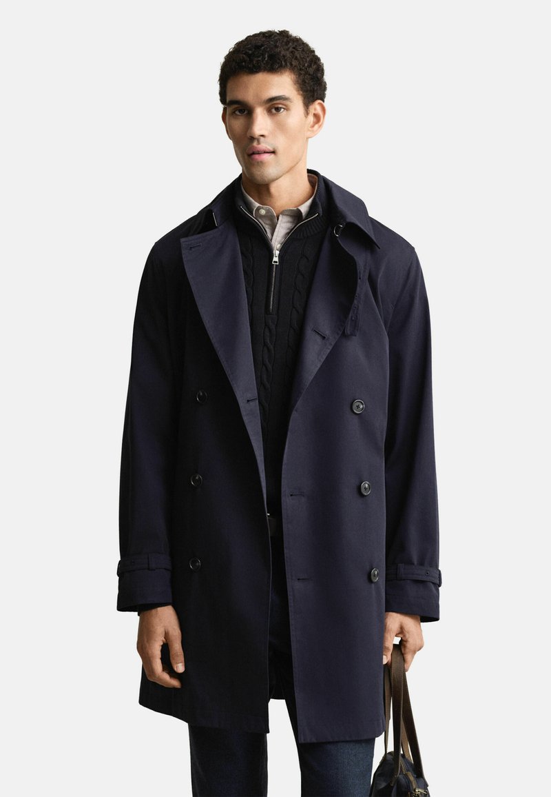 Granatowy, dwurzędowy płaszcz trench uszyty z gładkiego materiału, z szerokim kołnierzem, widocznymi guzikami i dopasowanym krojem, noszony na swetrze.