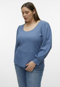 Lyseblå ribbet langærmet top med vid scoop-udskæring og puffede ærmer, parret med lyse denim jeans. Blød tekstur og afslappet pasform.