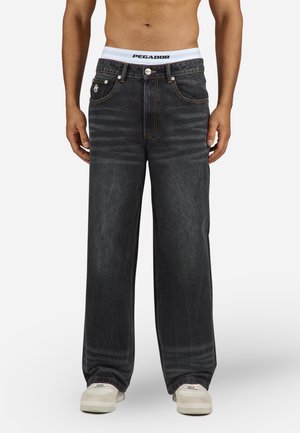 Brede zwarte denim jeans met oranje stiksels, voorzakken en een knoopsluiting; gedragen met een witte PEGADOR tailleband en crèmekleurige sneakers.