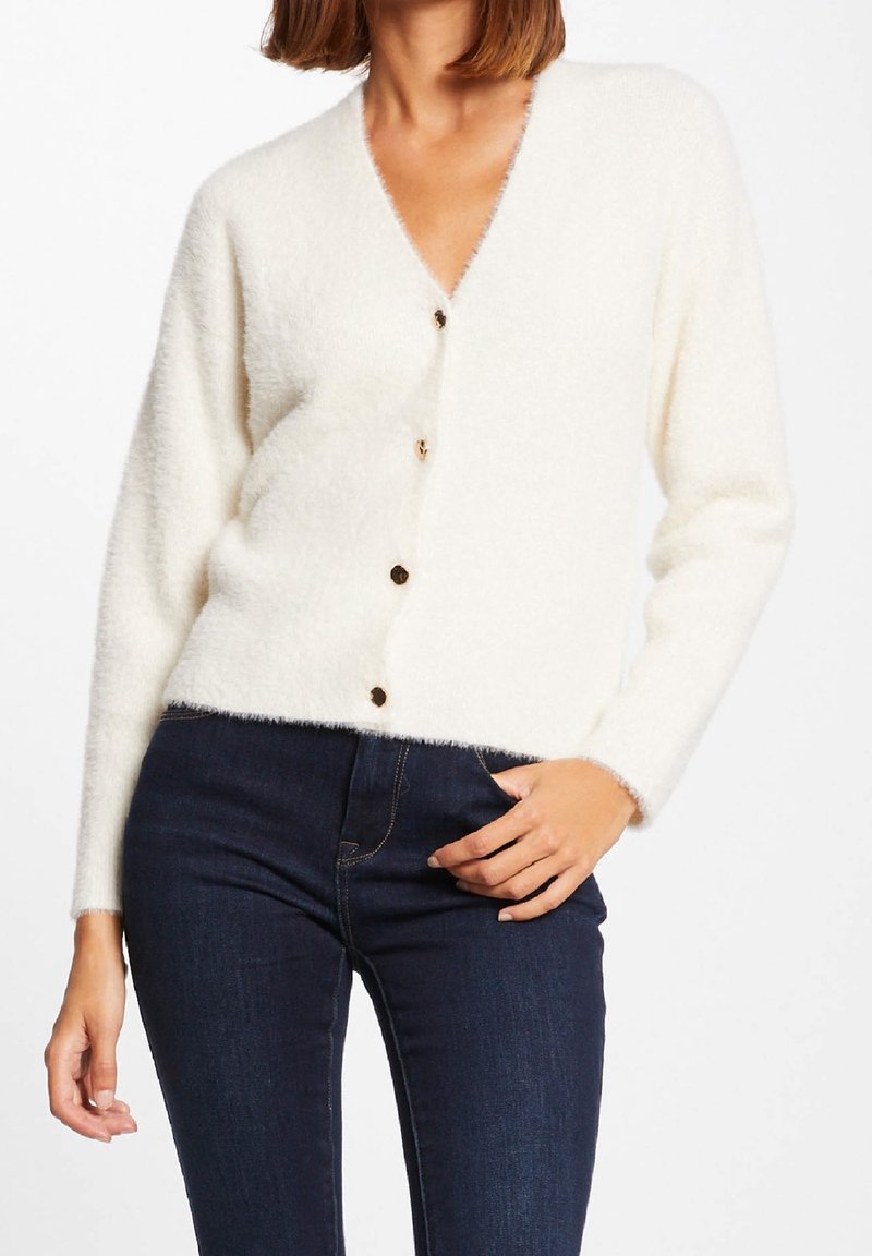 Femme portant un cardigan blanc duveteux boutonné avec manches longues et un jean skinny taille haute bleu foncé.