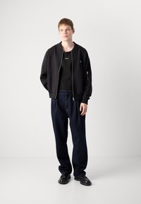 Ung mand står iført en sort lynlåsjakke, sort skjorte med hvid "HUGO" tekst, mørke løse jeans og sorte sko foran en hvid baggrund.