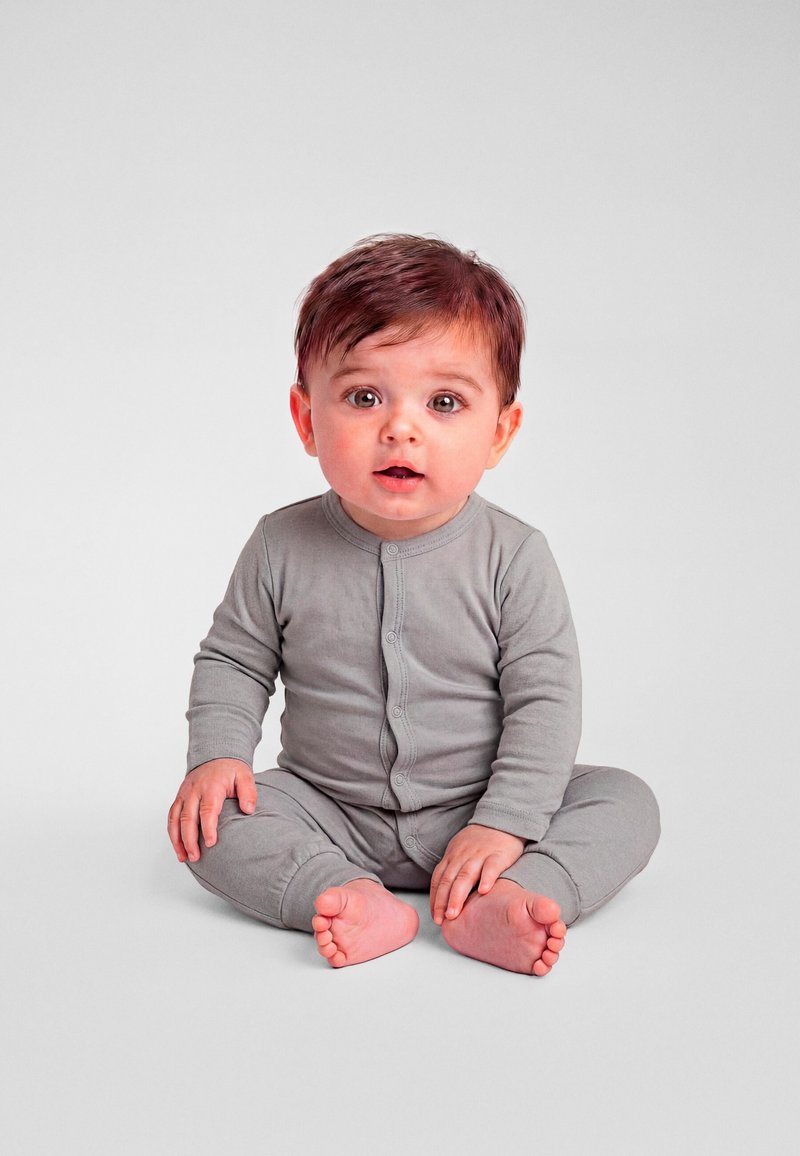 Tout-petit aux cheveux bruns assis en tailleur, portant une grenouillère grise boutonnée, regardant devant avec une expression neutre.