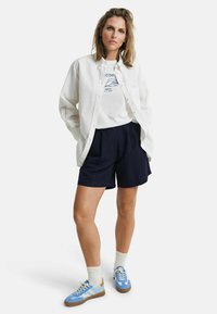 Chemise blanche à boutons sur un t-shirt graphique blanc, shorts navy et baskets bleues avec accents beiges. Cheveux courts, posture debout.