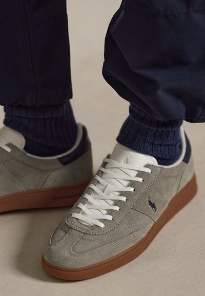 Light gray suede sneakers with white laces and brown soles worn with dark blue ribbed socks and black pants.