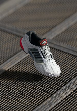 Zapato deportivo blanco con parte superior texturizada, acentos grises y logotipo rojo. Cuenta con un diseño de cordones y material liviano.