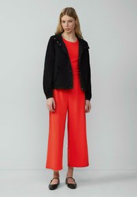Zwarte lichtgewicht jas met een capuchon, drukknoopsluitingen en voorzakken, gedragen over een felrode jumpsuit met brede broekspijpen. Zwarte ballerina's maken de look compleet.