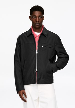 Veste noire avec fermeture éclair, col en velours côtelé et poche avant ; portée par-dessus une chemise rose à boutons. Pantalon blanc visible en bas.