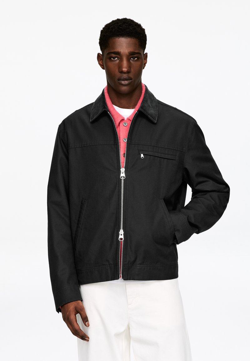 Giacca nera con chiusura a zip, colletto in velluto a coste e tasca anteriore; indossata sopra una camicia rosa con bottoni. Pantaloni bianchi visibili nella parte inferiore.