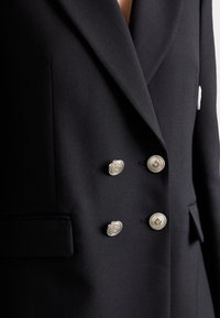 Blazer doppiopetto nero realizzato in tessuto testurizzato, con sei bottoni ornamentali in argento e una tasca sul petto.