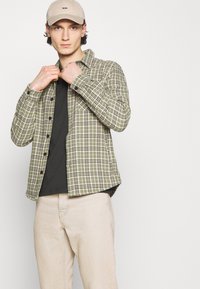 Grünes kariertes Flanellhemd mit Knopfleiste, getragen über einem schwarzen T-Shirt, kombiniert mit hellen Jeans und einer beigen Kappe.