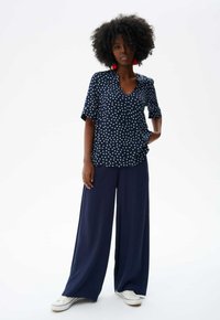 Marineblauwe blouse met polka dots en korte mouwen, gecombineerd met marineblauwe wijde broek en witte sneakers. De blouse heeft een V-hals en een casual pasvorm.