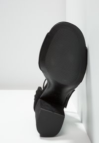 Chaussure à talon haut en daim noir avec un bout carré et une semelle extérieure en caoutchouc noir texturée. Comprend une bride en tissu autour de la cheville et une semelle intérieure rembourrée.