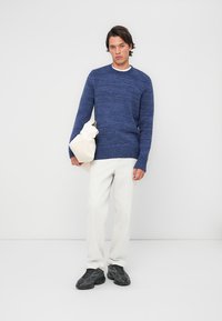 Modra pletena džemper s teksturiranim vzorcem, bela crewneck majica, v kombinaciji s smetanastimi hlačami in črnimi supergami, drži smetanasto torbico.