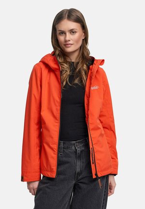 Kvinde iført en oplyst orange hættetrøje med åben lynlås over en sort skjorte og mørkegrå jeans, vendt fremad med afslappet udtryk.