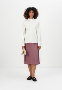 Top bianco a maniche lunghe con colletto alto, abbinato a una gonna rosa chiaro lunga fino al ginocchio. Scarpe nere senza lacci e borsa di paglia.